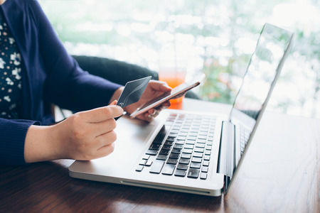 Young woman sitting in coffee shop at wooden table, drinking coffee and using smartphone.On table is laptop. Girl browsing internet, chatting, blogging. Female holding phone and looking on his screen.の写真素材