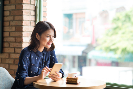 Beautiful cute asian young businesswoman in the cafe, using mobile phone and drinking coffee smilingの写真素材