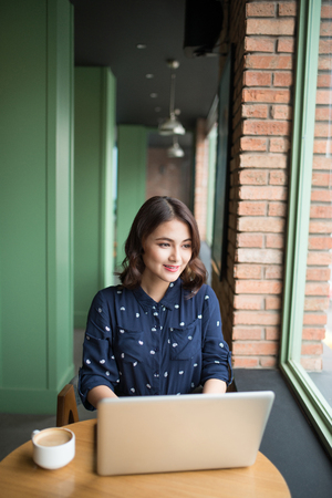 Beautiful cute asian young businesswoman in the cafe, using laptop and drinking coffee smilingの写真素材