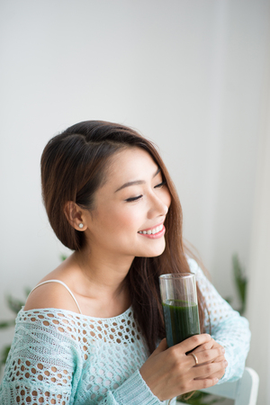 Smiling young asian woman drinking green fresh vegetable juice or smoothie from glass at homeの写真素材