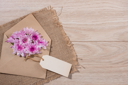 Blank white paper tag with brown envelope and pink flowers on wooden table.の写真素材