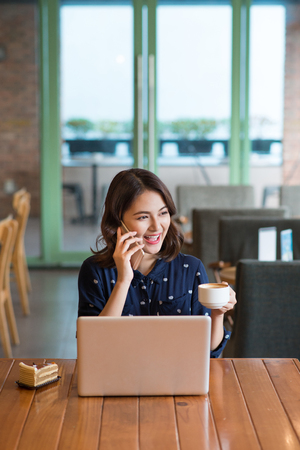 Beautiful cute asian young businesswoman in the cafe, listening to mobile phone and holding coffee mugの写真素材