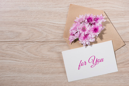 Blank white paper tag with brown envelope and pink flowers on wooden table.の写真素材