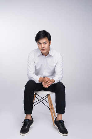 Smart casual asian man seated on chair, posing while looking at camera in studio backgroundの写真素材