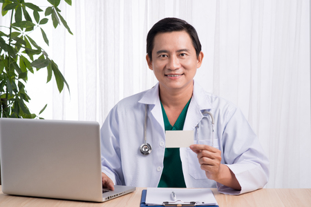 Smiling doctor sitting at his desk in medical office showing namecardの写真素材