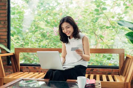 Confident young asian woman in smart casual wear shopping online while sitting near window in creative office or cafeの写真素材