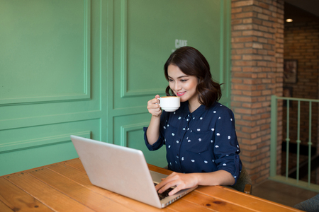 Beautiful cute asian young businesswoman in the cafe, using laptop and drinking coffee smilingの写真素材