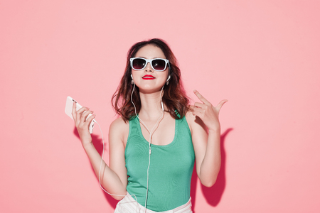 Young attractive asian woman with professional makeup singing and dancing while listening to music on pink background.の写真素材