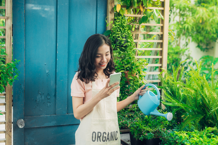 Young asian woman using smartphone, typing on phone while watering the plantsの写真素材