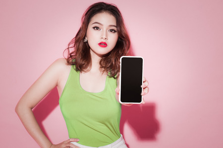 Asian girl showing smartphone screen on pink background.