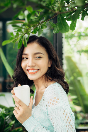Happy young asian woman with mug in hands drinking coffee standing on balcony.の写真素材