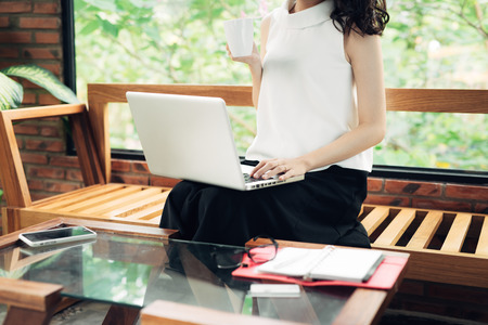 Confident young asian woman in smart casual wear shopping online while sitting near window in creative office or cafeの写真素材