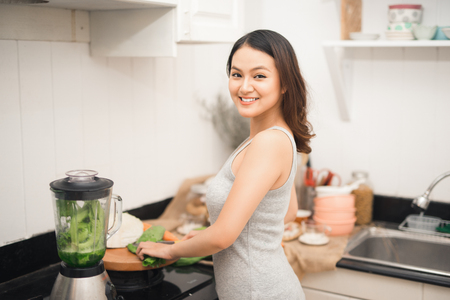 Smiling asian woman making smoothie with fresh vegetables in the blender in kitchen at home.の写真素材