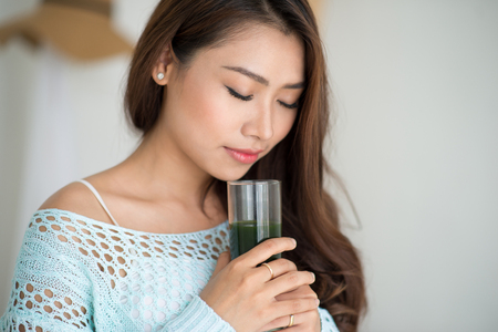 Pretty young asian woman drinking green fresh vegetable juice or smoothie from glass at homeの写真素材