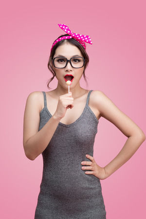 Portrait of beautiful asian woman eating heart shape lollipop, dressed and makeup in pin-up style on pink background.の写真素材