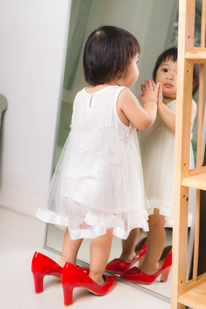Cute funny little baby girl walks at home trying mom's red high heel shoesの写真素材