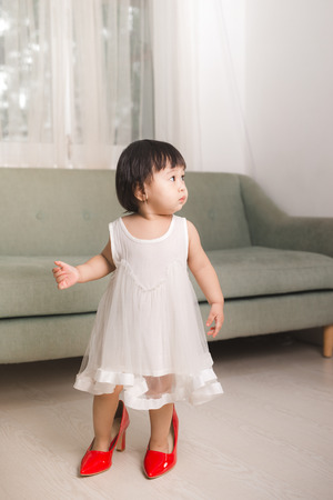 Cute funny little baby girl walks at home trying mom's red high heel shoesの写真素材