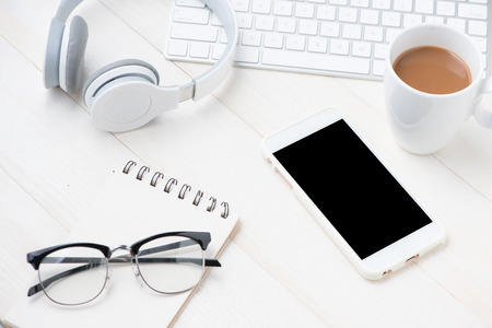 Notepad and computer keyboard on white table with mobile phone, headphone, eyeglasses, coffee cup. View from aboveの写真素材