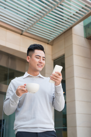 Handsome young man drinking coffee and using mobile phone at cityの写真素材