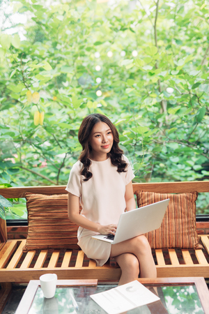 Beautiful young smiling woman working on laptop while sitting on bed at home near big window.の写真素材