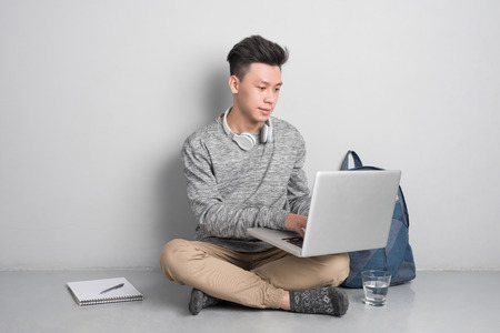 Young asian man in casual clothes is using a laptop, smiling while sitting on the floorの写真素材