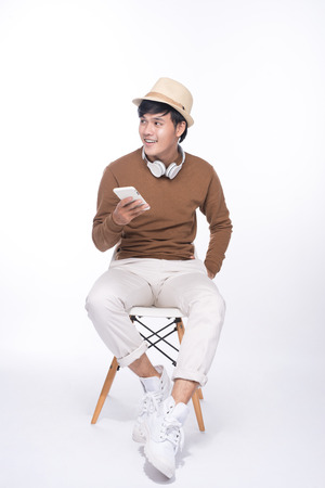 Smart casual asian man seated on chair, holding smartphone in studio backgroundの写真素材
