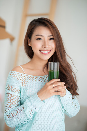 Pretty young asian woman drinking green fresh vegetable juice or smoothie from glass at homeの写真素材