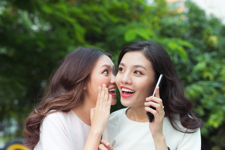 Surpried young woman whispering at a cheerful friend while on callの写真素材