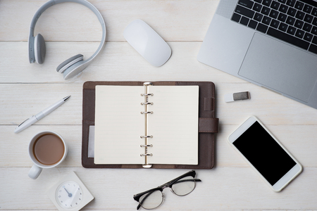 Top view on opened notebook, smartphone, laptop, eyeglasses, cup of coffee and other equipment on wooden office desk.の写真素材