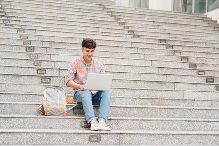 Handsome student in shirt working on laptop sitting on stairs in collegeの写真素材