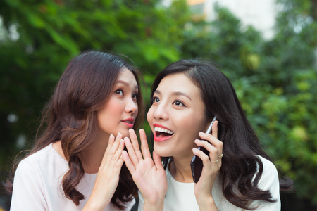 Young woman whispering into cheerful friend's ear while on callの写真素材