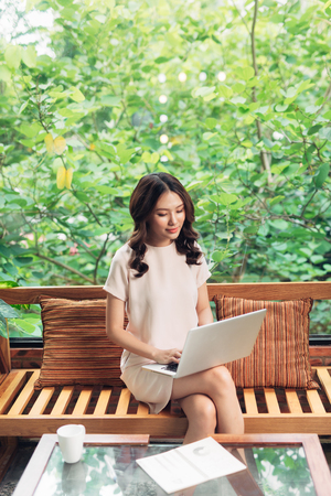 Beautiful young smiling woman working on laptop while sitting on bed at home near big window.の写真素材