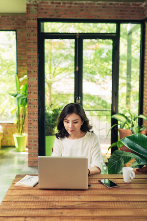 Beautiful young smiling woman working on laptop while sitting on bed at home near big window.の写真素材