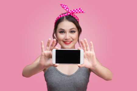 Beautiful young asian woman with pin-up make-up and hairstyle over pink background with mobile phoneの写真素材