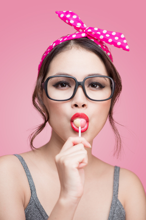 Portrait of beautiful asian woman eating heart shape lollipop, dressed and makeup in pin-up style on pink background.の写真素材