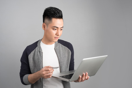Young asian man with laptop and credit card in studio isolated gray backgroundの写真素材