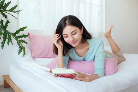 Young asian woman lying on bed, reading bookの写真素材