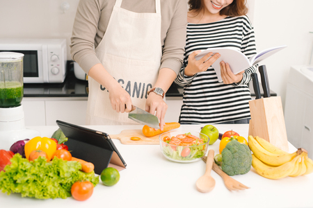 Happy couple in kitchen together. Man cutting vegetables and making salad while his wife reading recipeの写真素材