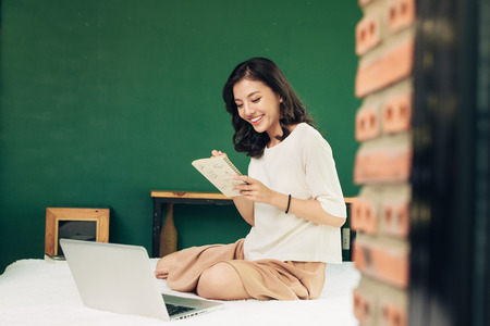 Beautiful young smiling woman working on laptop while sitting on bed at homeの写真素材