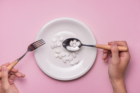 Top view of man's hands at dining table holding a fork and knife above dish with pills over pink background.の写真素材