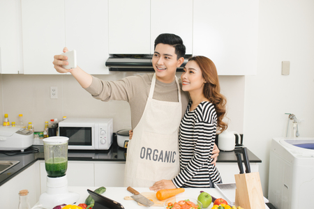 Happy asian couple smiling and cooking in the kitchen doing selfie using a smart phoneの写真素材