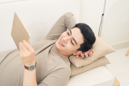 Enjoying time at home. Cheerful young man holding digital tablet and looking relaxed while lying on the couch at homeの写真素材