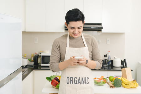 Handsome asian man texting with his smartphone in kitchenの写真素材