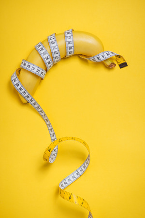 Diet, fitness and health concept presented by yellow banana wrapped in measure tape isolated on yellow paper backgroundの写真素材