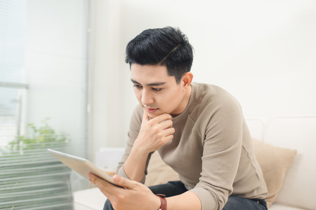 Handsome man is using a digital tablet and smiling while resting on couch at homeの写真素材