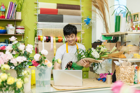 Happy asian male florist using laptop at counter in flower shopの写真素材