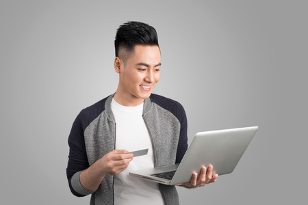 Young asian man with laptop and credit card in studio isolated gray backgroundの写真素材