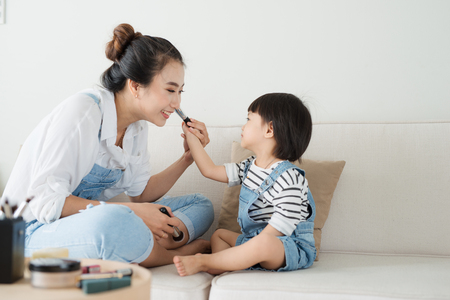 Cute little girl and her beautiful mother are doing makeup while sitting on couch at homeの写真素材