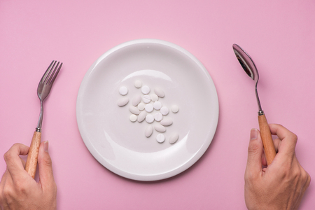 Top view of man's hands at dining table holding a fork and knife above dish with pills over pink background.の写真素材