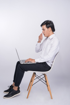 Smart casual asian man seated on chair, using laptop in studio backgroundの写真素材